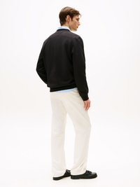 Schwarzer Sweatshirt, hellblaues Hemd darunter, weiße Hose und schwarze Schuhe, von hinten betrachtet, minimalistischer Hintergrund.