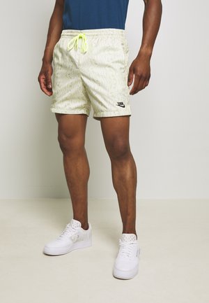 Mand iført lysegule mønstrede Nike shorts med sort logo, hvide sneakers og en mørkeblå skjorte, stående på en lys gulv- og vægbaggrund.