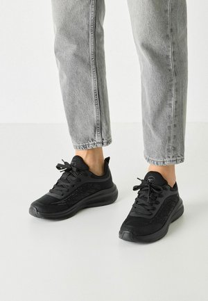 Baskets de sport noires avec un dessus en maille texturée, des lacets plats, un col rembourré et une semelle en caoutchouc épaisse. Portées avec un jean gris retroussé.