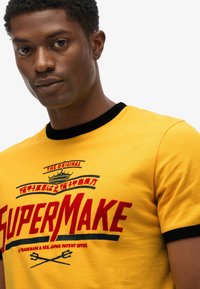 Yellow cotton T-shirt featuring black ribbed collar, bold red and black graphic text, and a crown design on the upper front.