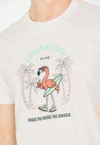 Vit t-shirt i bomull med ett grafiskt motiv av en pink flamingo som bär solglasögon och håller i en grön surfbräda, med palmer i bakgrunden.