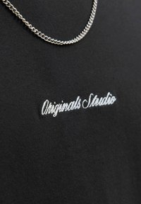 Musta t-särk, millel on valgest tikandist kiri "Originals Studio" ja hõbedane ketipaar, mis on kaela ümber asetatud.