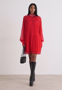 Crvena plisirana haljina s dugim prozirnim rukavima i visokim ovratnikom; model drži malu crnu torbicu; uparena s crnim čizmama do koljena.