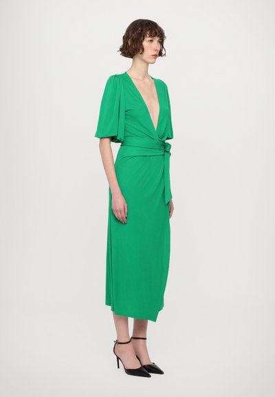 Green wrap dress with a deep V-neck, short sleeves, and a tied waist detail, paired with black strappy high heels. Smooth texture.