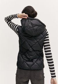 Chaleco acolchado negro con capucha, con un diseño de diamantes. La persona lleva puesta una camisa de rayas negras y blancas de manga larga y pantalones grises.
