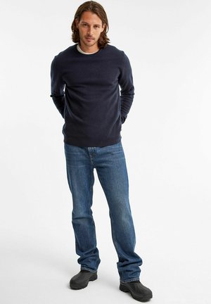 Marineblauer Rundhals-Pullover, leichtes Material, zeitloses Design. Kombiniert mit blauen Jeans, lockerer Passform und schwarzem Schuhwerk. Einfach und funktional.