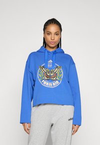 Žena s pletenom kosom, odjevena u plavu kapu s grafikonom leptira i logotipom "Farm Rio", te svijetlosivim Adidas trenirkama, stoji nasuprot jednostavne pozadine.