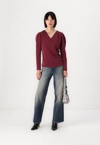 Burgunder fôret bluse med puff-sleeve, v-halsdesign; kombinert med lyse, vide jeans; modellen bærer en liten metallisk håndveske.