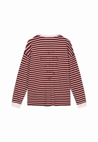 Marškiniai ilgomis rankovėmis bordo ir grietinėlės spalvų horizontalios juostelės su balta apykakle. Tekstas ant nugaros sako: "RAŠAU SAVO KURIAMĄ MAGIJĄ."