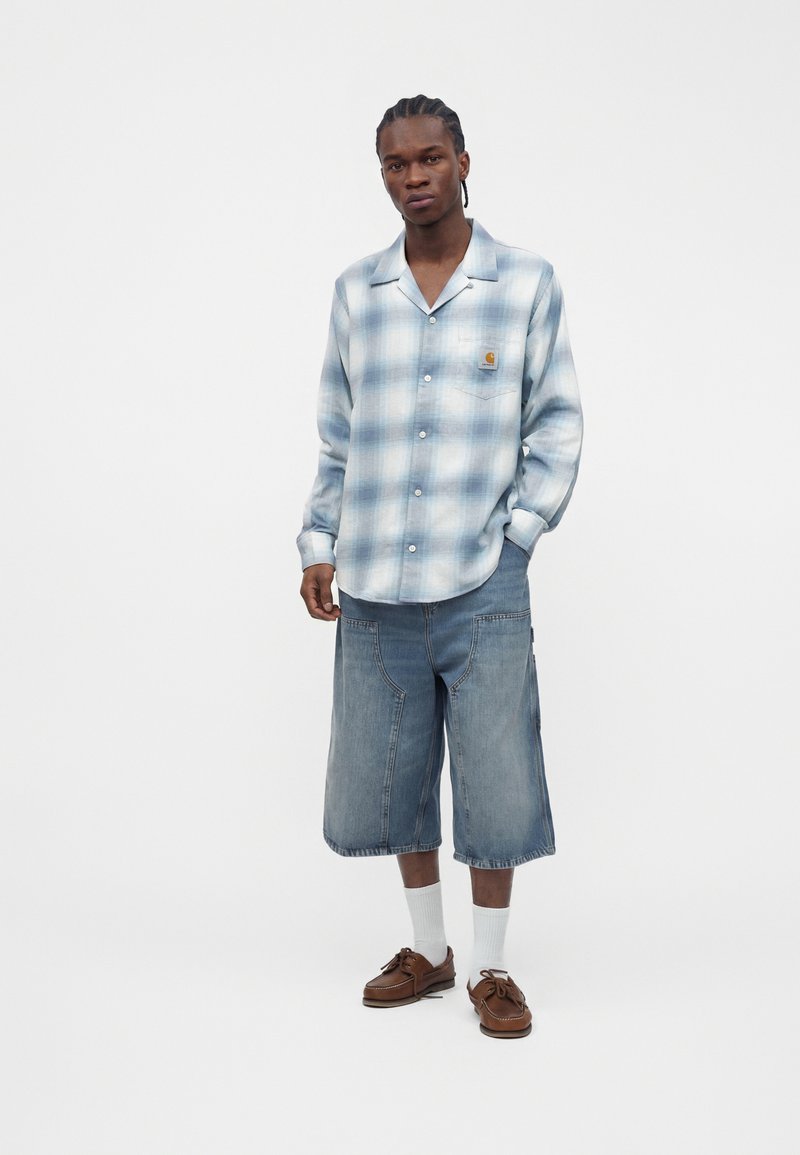 Man wearing light blue plaid button-up shirt, long denim shorts, white socks, and brown lace-up shoes, standing with one hand in pocket.