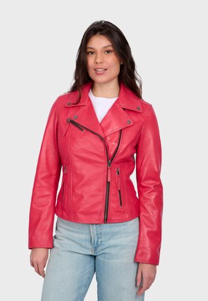 Mladá žena v jasně růžové kožené motorkářské bundě se zipy a světlých modrých džínách, stojící na světle šedém pozadí.