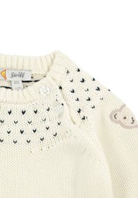 Maglione lavorato a maglia crema con motivi a triangolo neri, due bottoni sulla spalla e un'appliqué di un orso beige sul petto. Tag della taglia: 62.