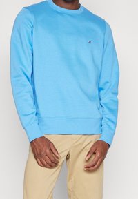 Světle modrý crewneck svetr vyrobený z měkké směsi bavlny. Obsahuje dlouhé rukávy, žebrované manžety a malé logo na hrudi.