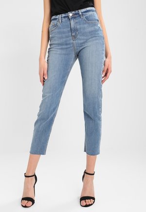 Femme portant un jean court bleu clair avec des ourlets effilochés et des sandales noires à talons avec une bride à la cheville, debout devant un fond uni.