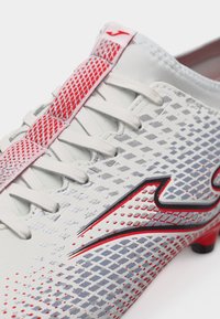 Λευκό ποδοσφαιρικό παπούτσι με κόκκινα και γκρι γεωμετρικά σχέδια, λευκά κορδόνια και κόκκινο τσόφλι τράβηγμα στη γλώσσα.