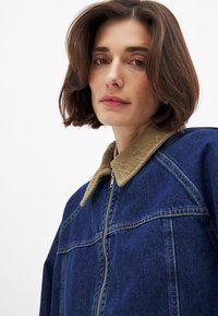 Tumši zila denim jaka ar korduroja apkakli. Iespējas ietver pilnu rāvējslēdzēju un izšūtas plecu šuves, kas parāda brīvu piegriezumu.