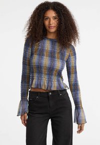 Blauwe en gele gestreepte lange mouw top met een smockdesign en uitlopende manchetten, gecombineerd met zwarte jeans. De stof lijkt lichtgewicht en textured.
