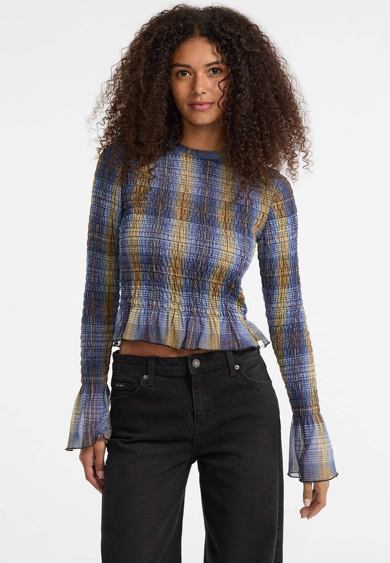 Blauwe en gele gestreepte lange mouw top met een smockdesign en uitlopende manchetten, gecombineerd met zwarte jeans. De stof lijkt lichtgewicht en textured.