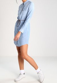 Světle modré džínsové šaty košilového střihu s dlouhými rukávy, zapínání na knoflíky, dvěma náprsními kapsami a přiléhavým pasem, doplněné bílými teniskami.