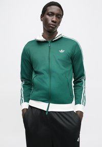 Green zip-up jacket made of smooth fabric with white accents, ribbed cuffs, and a small logo on the chest; worn with black pants.