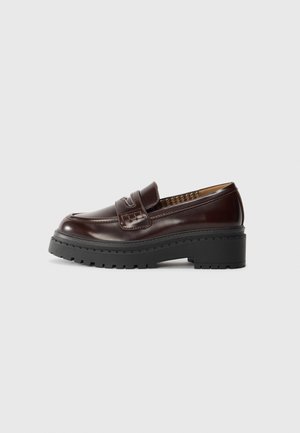 Mocassino in pelle marrone scuro con suola robusta nera a tasselli e dettaglio della cinturini cuciti, mostrato di profilo su sfondo bianco.