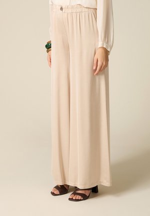 Pantaloni beige chiaro a gamba larga con vita elastica e dettaglio con bottoni, indossati con sandali neri con tacco e punta aperta e braccialetti verdi.