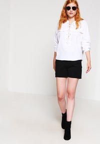 Blusa bianca a maniche lunghe con laccio frontale, abbinata a pantaloni corti neri e stivali neri alla caviglia. Modella con occhiali da sole. Tessuto morbido.