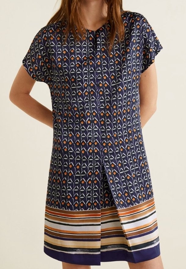 Femme portant une robe bleu marine avec des motifs géométriques orange et blancs et des rayures horizontales près de l'ourlet, debout les mains derrière le dos.