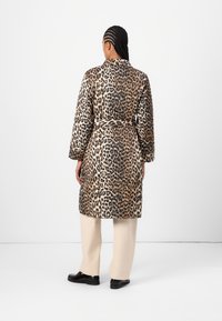 Femme aux cheveux tressés portant un manteau mi-long imprimé léopard, un pantalon beige et des chaussures noires, debout devant un fond blanc uni.