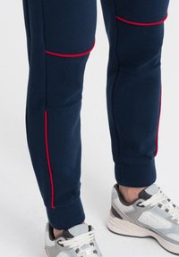 Marintröjor med röda accenter, med ribbade texturer på knäna och ärmsluten. Bärs med grå sneakers som betonar den sportiga designen och komforten.