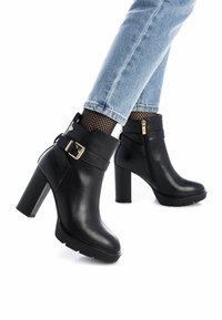 Botas negras de cuero hasta el tobillo con un tacón grueso, que cuentan con un cierre lateral y una correa con una hebilla dorada, situadas sobre unos jeans azules y medias de rejilla.