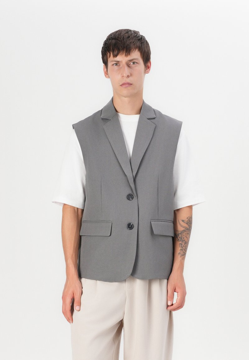Šedý bezrukávový blazer s dvoupolohovým zapínáním a předními kapsami, vyrobený z hladkého materiálu, nošený přes bílé tričko a béžové kalhoty.