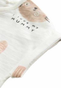 Monedero de algodón blanco con mangas cortas, que presenta un gráfico de una cara de animal marrón y el texto "Amo a mi MAMÁ". Cierre con botones a presión en el hombro.