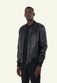 Zwarte leren bomberjack met ribgebreide manchetten en boord, sluiting met rits aan de voorkant, zijzakken en een klein borstzakje. Gladde textuur.