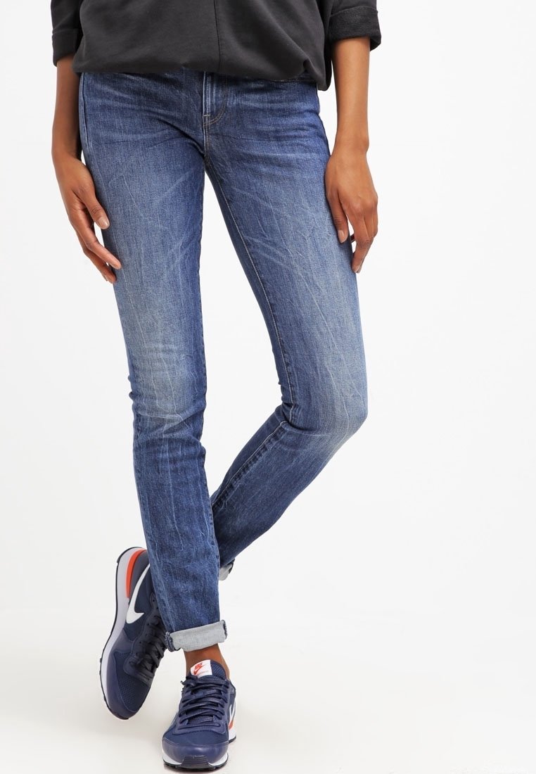 Persona con jeans azules remangados y zapatillas Nike azul marino, de pie con una pierna cruzada sobre la otra.