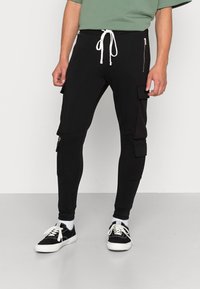 Zwarte cargo broek in een getailleerd ontwerp met een witte trekkoord, zijzakken met rits en een gestructureerde stof. Gecombineerd met zwarte sneakers.