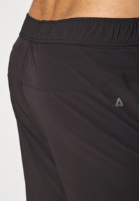 Detail černých sportovních šortek s elastickým pasem na osobě, zobrazující texturu látky a malý trojúhelníkový logo na boku.