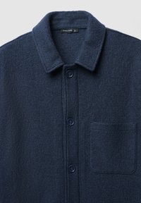 Giacca camicia in lana blu navy con colletto classico, caratterizzata da una tasca anteriore e bottoni tono su tono. Tessuto strutturato con finitura morbida.