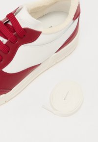 Piros és fehér bőr sneaker, kerek orral, texturált piros panelekkel és piros fűzőkkel. A cipő mellett egy tekeredett fehér fűző található.