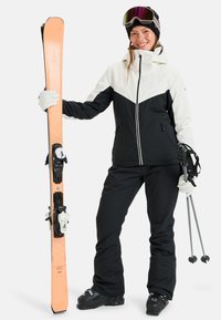 Kurtka narciarska w czarno-białym kolorze z wzorem w zygzak, zestawiona z czarnymi spodniami. Narciarz trzyma pomarańczowe narty z czarnymi wiązaniami i kijami.