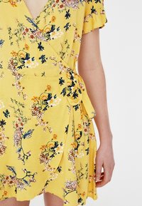 Robe portefeuille jaune avec un motif floral aux accents bleus et orange. Manches courtes, encolure en V et tissu texturé.