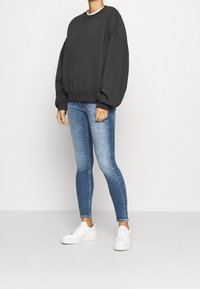 Sudadera gris oscuro de gran tamaño con hombros caídos, combinada con jeans ajustados azules. Zapatillas blancas y una blusa blanca de varias capas visible debajo.
