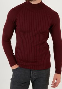 Burgunderfarbener, gerippter Langarm-Pullover mit hohem Kragen und tailliertem Schnitt, aus einem strukturierten Strickmaterial gefertigt.