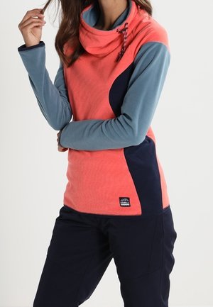 Kvinde iført en koral- og marineblå pullover med grå ærmer, med en høj krave med snore, kombineret med sorte bukser.