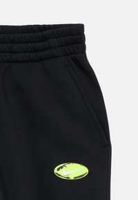 Calças de treino pretas com uma cinta elástica e bolsos laterais, apresentando um destacado logótipo da Nike em verde-neon na coxa esquerda.