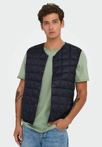 Gilet matelassé bleu marine avec boutons-pression, porté par-dessus un t-shirt vert clair. Présente un design minimaliste et un tissu doux et texturé.