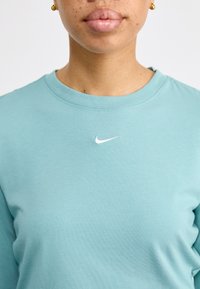 Šviesiai mėlynas medvilninis marškinėlis su apvalia apykakle, su mažu balto Nike logotipu, esančiu centre ant krūtinės. Paprasta, lygi audinio tekstūra.