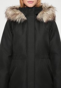 Kobieta w czarnym zimowym płaszczu z wysokim kołnierzem i beżowym kapturem wykończonym futrem, pokazana od szyi do talii.