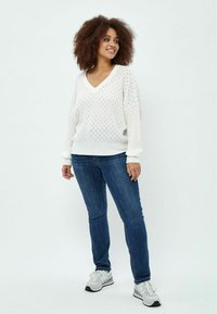 Bela pletena jopica z V-izrezom in oversized rokavi, v kombinaciji s temno modrimi skinny kavbojkami in svetlo sivimi teniskami.