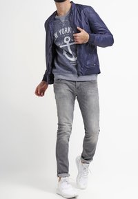 Marinblå bombarjacka av slätt tyg över en grå sweatshirt med en ankarsgrafik, kombinerad med ljusa jeans och vita sneakers.